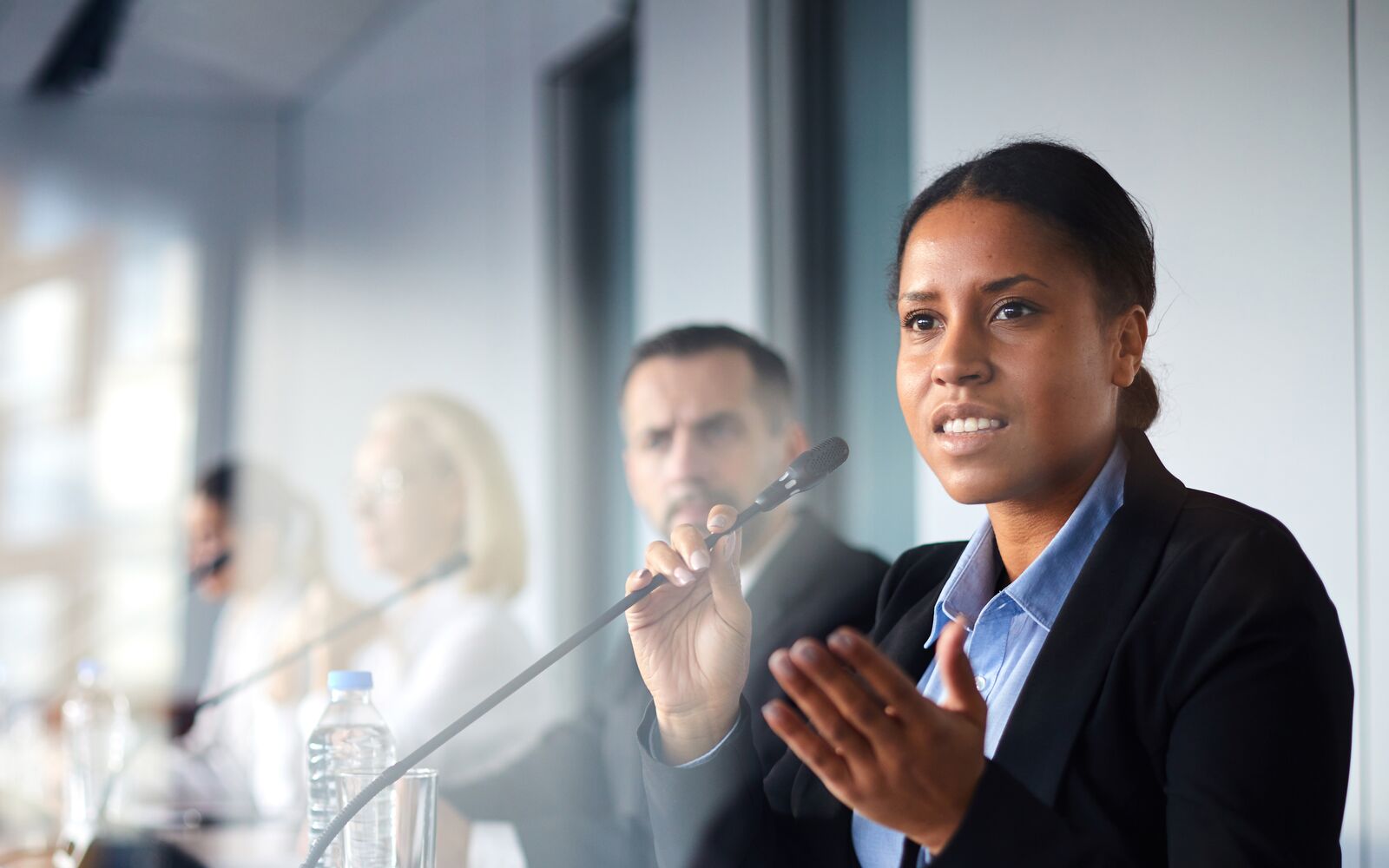 Young female deputee in elegant suit sitting at conference and speaking in microphone to colleague; Shutterstock ID 1312349519; purchase_order: -; job: -; client: -; other: -